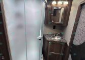 Compact RV bathroom with curved glass shower, skylight, and wooden vanity with sink and mirrored medicine cabinet lighting.