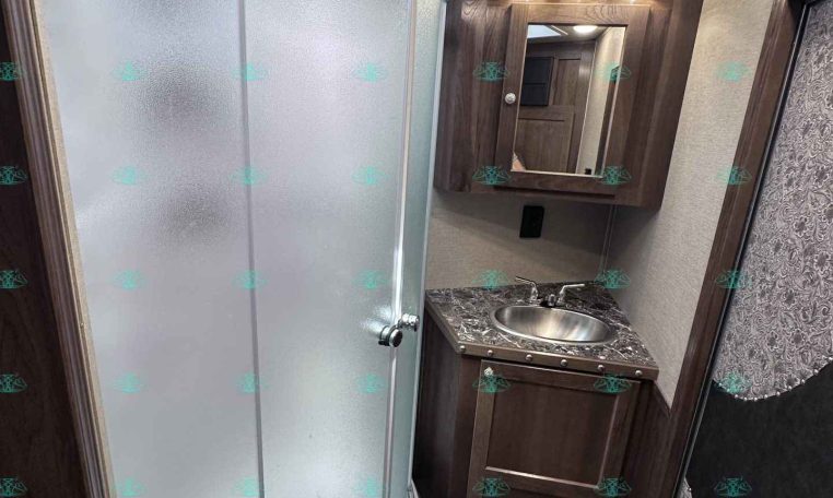 Compact RV bathroom with curved glass shower, skylight, and wooden vanity with sink and mirrored medicine cabinet lighting.