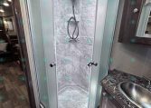 Curved glass shower enclosure in an RV bathroom with tiled walls, skylight, and a nearby sink with a dark wood vanity edge.