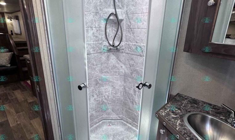 Curved glass shower enclosure in an RV bathroom with tiled walls, skylight, and a nearby sink with a dark wood vanity edge.