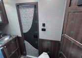 Compact RV bathroom with a toilet in foreground, dark wood cabinets, and a sink to the left; doorway with decorative panel ahead.