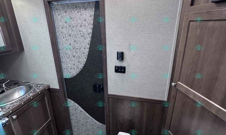 Compact RV bathroom with a toilet in foreground, dark wood cabinets, and a sink to the left; doorway with decorative panel ahead.