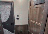 RV bathroom with wood-paneled walls, white toilet in foreground, and a curved glass shower with patterned curtain on the left; skylight above.
