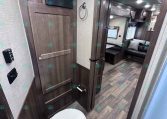Interior shot of an RV bathroom with dark wood cabinets, a toilet in the foreground, and a doorway leading to a living area with a sofa visible in the background.