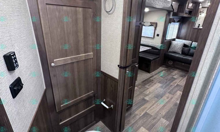 Interior shot of an RV bathroom with dark wood cabinets, a toilet in the foreground, and a doorway leading to a living area with a sofa visible in the background.