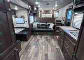 Interior of a modern travel trailer: kitchen with dark wood cabinets, sofa and dinette, and a rear sleeping area.