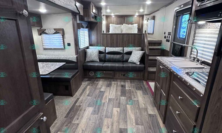 Interior of a modern travel trailer: kitchen with dark wood cabinets, sofa and dinette, and a rear sleeping area.
