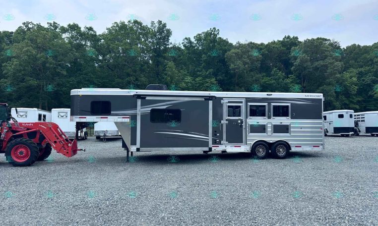 Silver and gray horse trailer on a gravel lot, with a red tractor in the foreground and several trailers beside it, trees and a blue sky in the background. Bottom edge shows decorative text reading CarolinaTrailersSales.com.