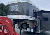 Dark gray Merhow Stampede horse trailer with silver trim, open side door, and attached red farming equipment in a gravel lot.