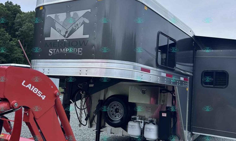 Dark gray Merhow Stampede horse trailer with silver trim, open side door, and attached red farming equipment in a gravel lot.