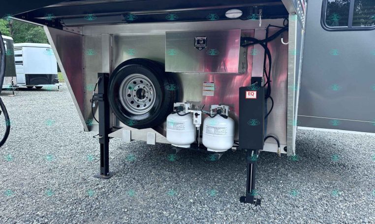 Open storage compartment of a trailer showing a spare tire, propane tanks, and a metal toolbox with cables and hoses inside a metallic enclosure.