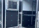 Open trailer rear doors reveal a dark interior with a metal bulkhead, a small barred window, and a silver loading ramp outside on gravel.
