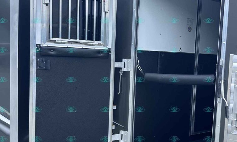 Open trailer rear doors reveal a dark interior with a metal bulkhead, a small barred window, and a silver loading ramp outside on gravel.