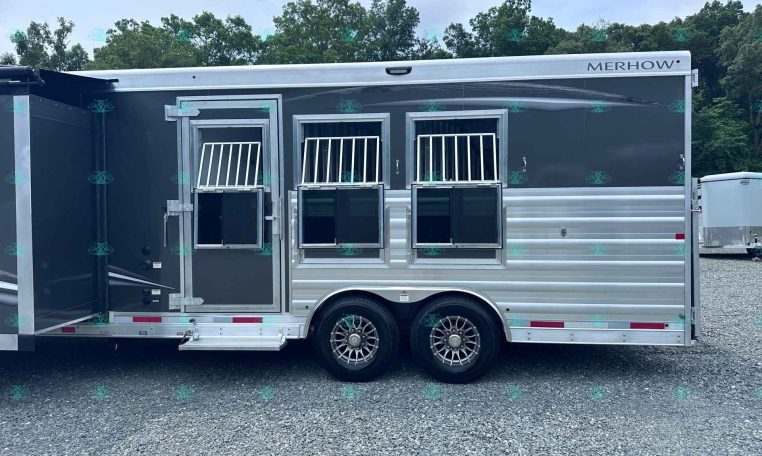 Side view of a dark gray Merhow horse trailer with three barred windows and tandem wheels parked on gravel.