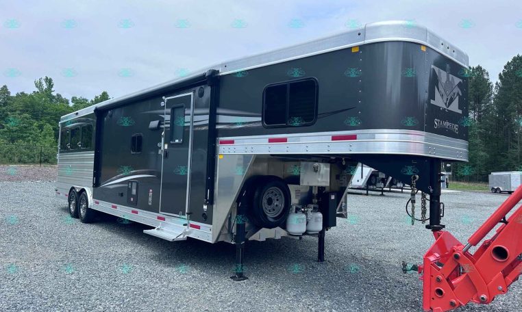 Large dark gray gooseneck horse trailer with living quarters, parked on a gravel lot with a side door and windows visible, propane tanks beneath.”