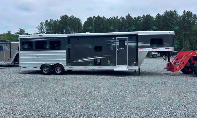 Side view of a black and silver Merhow horse trailer with living quarters, parked on a gravel lot with trees behind.