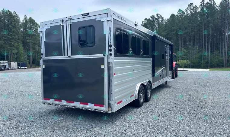 Silver-and-black Merhow horse trailer parked on a gravel lot with a forest backdrop; rear and side doors visible.