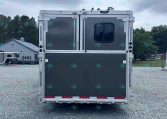 Rear view of a silver trailer with a small window, double doors, and red reflectors on a gravel lot, with trees and a gray building in the background under a cloudy sky.