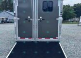 Back of a gray cargo trailer with double doors and a lowered ramp on a gravel lot; trees and a building visible in the distance.