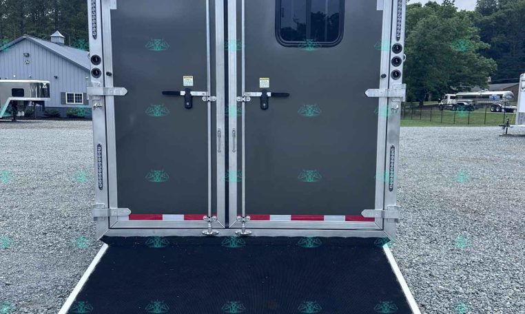Back of a gray cargo trailer with double doors and a lowered ramp on a gravel lot; trees and a building visible in the distance.