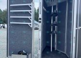 Open aluminum trailer with a dark, textured interior and a lowered, black ramp outside the doorway; tools and racks along the walls are visible inside.