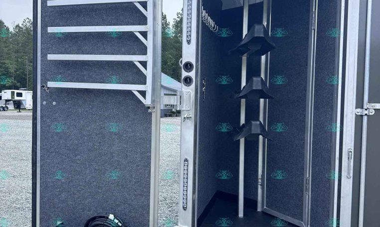 Open aluminum trailer with a dark, textured interior and a lowered, black ramp outside the doorway; tools and racks along the walls are visible inside.