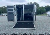 Open aluminum trailer with its doors swung wide, showing a dark interior with a vertical rack and shelving along the left wall, and a ramp lowered to the gravel ground in a trailer lot with other trailers in the background.