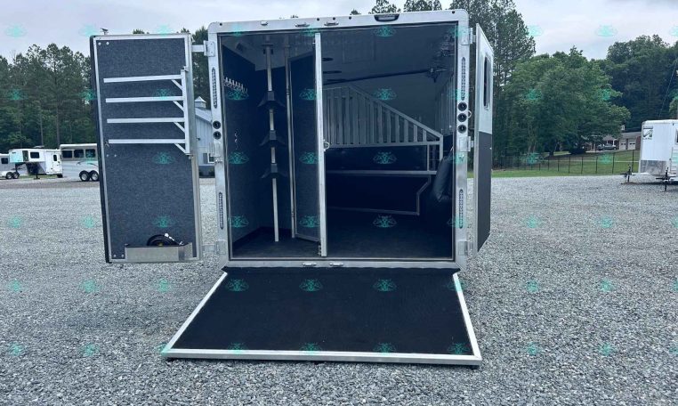 Open aluminum trailer with its doors swung wide, showing a dark interior with a vertical rack and shelving along the left wall, and a ramp lowered to the gravel ground in a trailer lot with other trailers in the background.