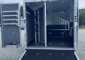 Open horse trailer interior with a divider, tack hooks on the left wall, and a lowered ramp onto a gravel surface.