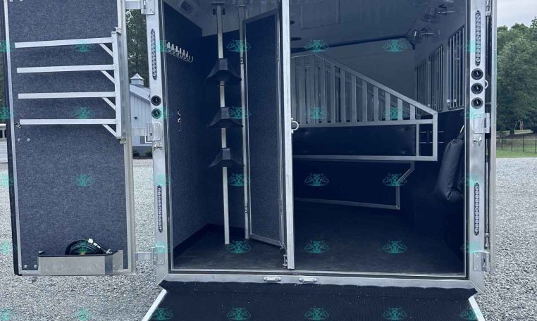 Open horse trailer interior with a divider, tack hooks on the left wall, and a lowered ramp onto a gravel surface.