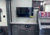 Interior of a modern trailer with a mounted TV above dark wood cabinets and a bunk bed to the right of the TV wall.