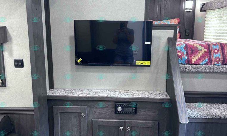 Interior of a modern trailer with a mounted TV above dark wood cabinets and a bunk bed to the right of the TV wall.