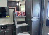 Dark wood RV interior with stairs to an elevated bed, colorful quilt, and built-in cabinets; TV on the left and entry door on the right.