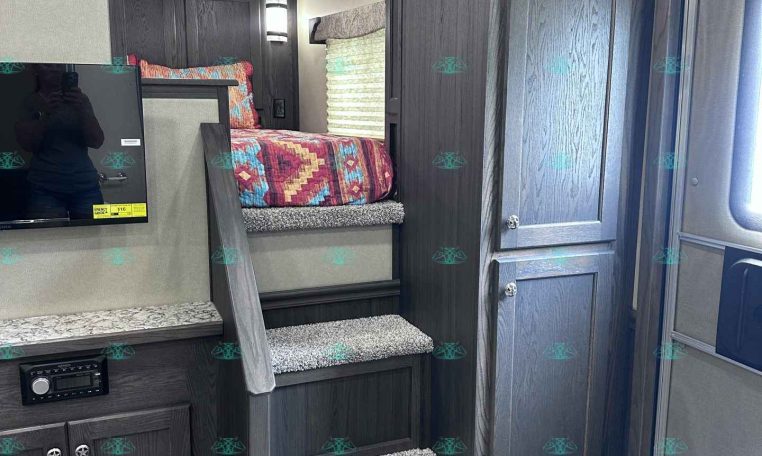 Dark wood RV interior with stairs to an elevated bed, colorful quilt, and built-in cabinets; TV on the left and entry door on the right.