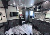 Interior view of a modern RV kitchen: dark wood cabinets, stainless microwave, countertop stove, sink, and a mounted TV opposite a small seating area with patterned pillows. A compact living area inside a travel trailer with stairs, cabinetry, and a windowed door at the back.