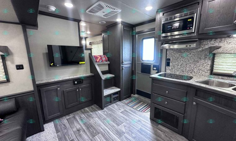 Interior view of a modern RV kitchen: dark wood cabinets, stainless microwave, countertop stove, sink, and a mounted TV opposite a small seating area with patterned pillows. A compact living area inside a travel trailer with stairs, cabinetry, and a windowed door at the back.