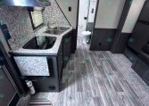 Compact trailer kitchen with granite counters, double sink, stove, and mounted microwave; doorway to bathroom in the back.