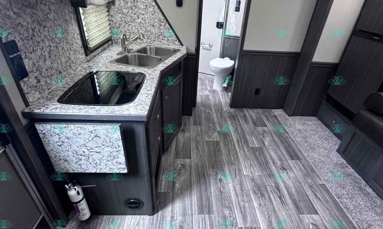Compact trailer kitchen with granite counters, double sink, stove, and mounted microwave; doorway to bathroom in the back.