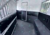 Interior of a white horse trailer with a central divider, black padded lower walls, and metal rails along the left side; barred window on the right and a small access door in the back area