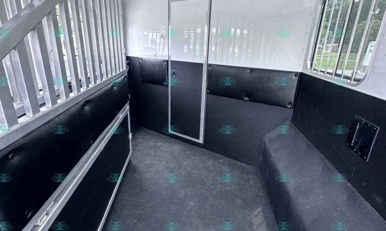 Interior of a white horse trailer with a central divider, black padded lower walls, and metal rails along the left side; barred window on the right and a small access door in the back area