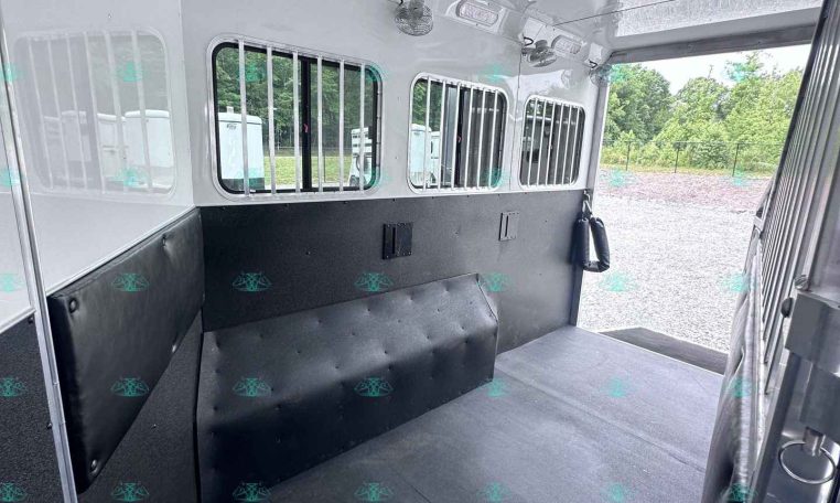 Interior of a white horse trailer with padded black walls, barred windows, ceiling lights, and ceiling fans visible.