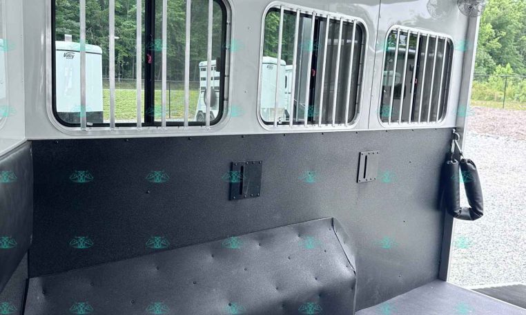 Interior of a horse trailer showing barred stalls, white walls, ceiling fans, and a dark padded lower wall bar.