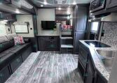 Interior of a modern RV living area: gray sofa with patterned pillows, TV wall, and kitchenette on the right with a double sink and microwave above.