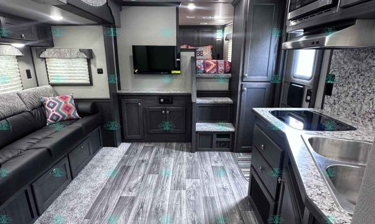 Interior of a modern RV living area: gray sofa with patterned pillows, TV wall, and kitchenette on the right with a double sink and microwave above.