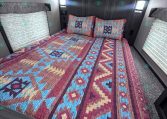 Cozy trailer bedroom with a colorful geometric quilt, two pillows, and dark wood headboard against paneled walls. Blinds cover side windows, ceiling lights visible.