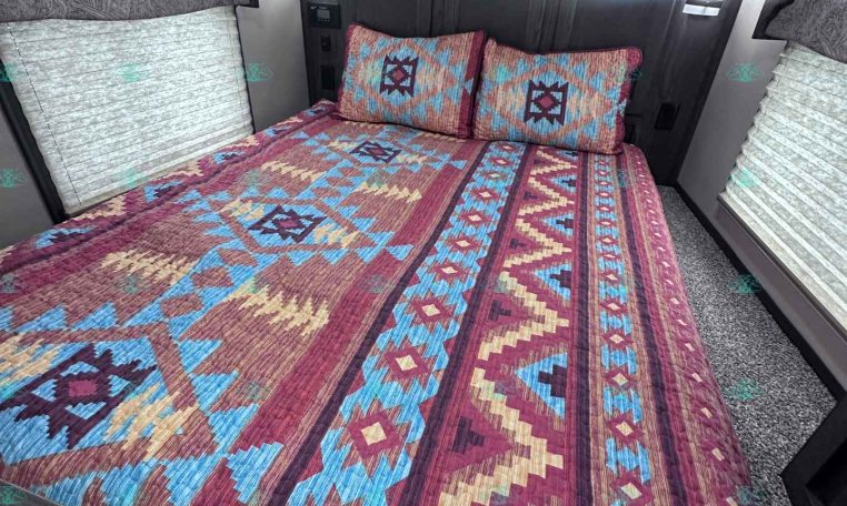 Cozy trailer bedroom with a colorful geometric quilt, two pillows, and dark wood headboard against paneled walls. Blinds cover side windows, ceiling lights visible.