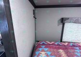 Bedroom in a travel trailer with a colorful geometric quilt on the bed, beige walls, and dark wood trim around the door and window shade.