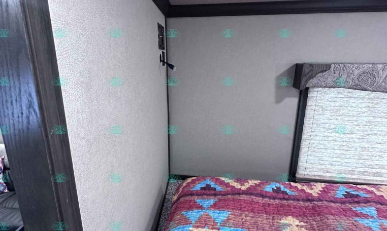 Bedroom in a travel trailer with a colorful geometric quilt on the bed, beige walls, and dark wood trim around the door and window shade.