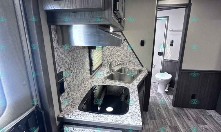 Compact RV kitchen with granite countertop, double sink, and a stove topped by a microwave/hood unit above. Nearby is a doorway to a bathroom.
