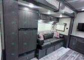 Modern RV interior with dark wood cabinetry, gray sofa with colorful patterned pillows, and overhead storage; built-in TV area and kitchen cabinets run along the wall.